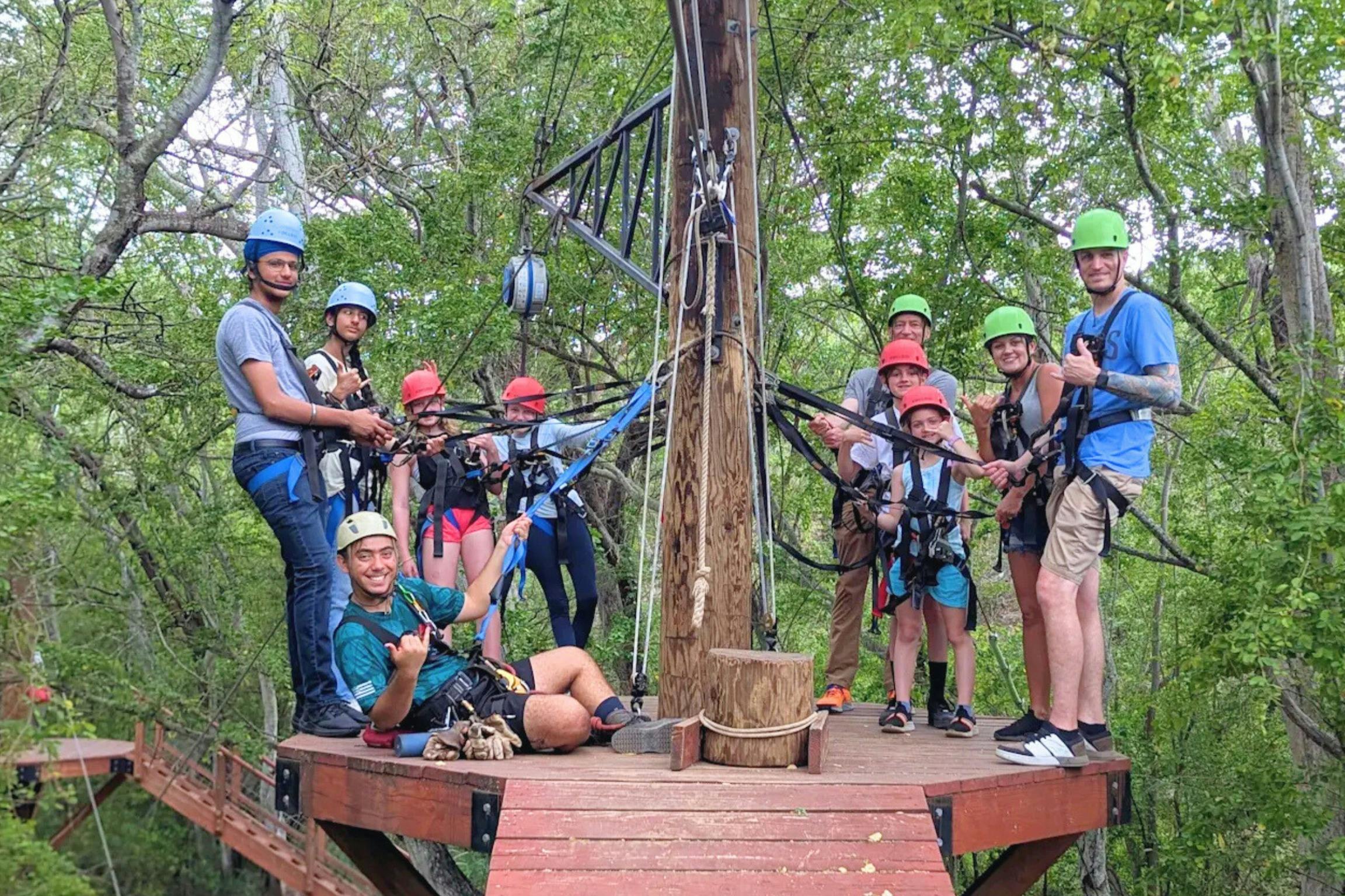 En gruppe mennesker med hjelme og seler, der står på en zipline-platform omgivet af træer og smiler efter et billede.
