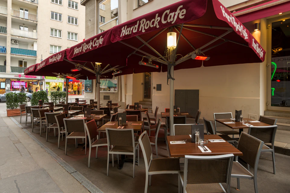Outdoor seating area of a Hard Rock Cafe with maroon umbrellas, tables, and chairs. Surrounding buildings are visible.