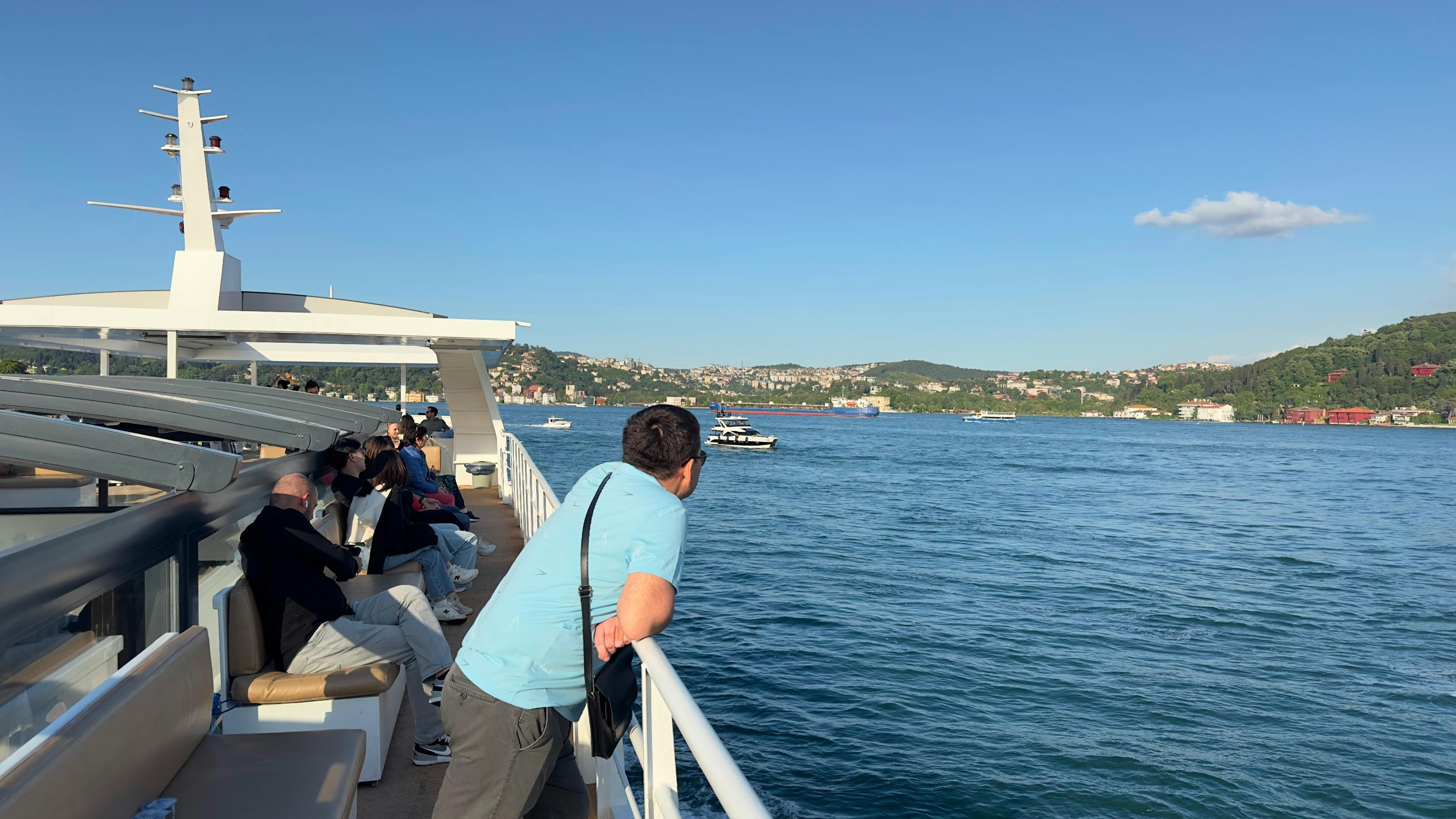 People on a ferry enjoying the view of a calm body of water with hills and buildings in the distance under a clear sky.