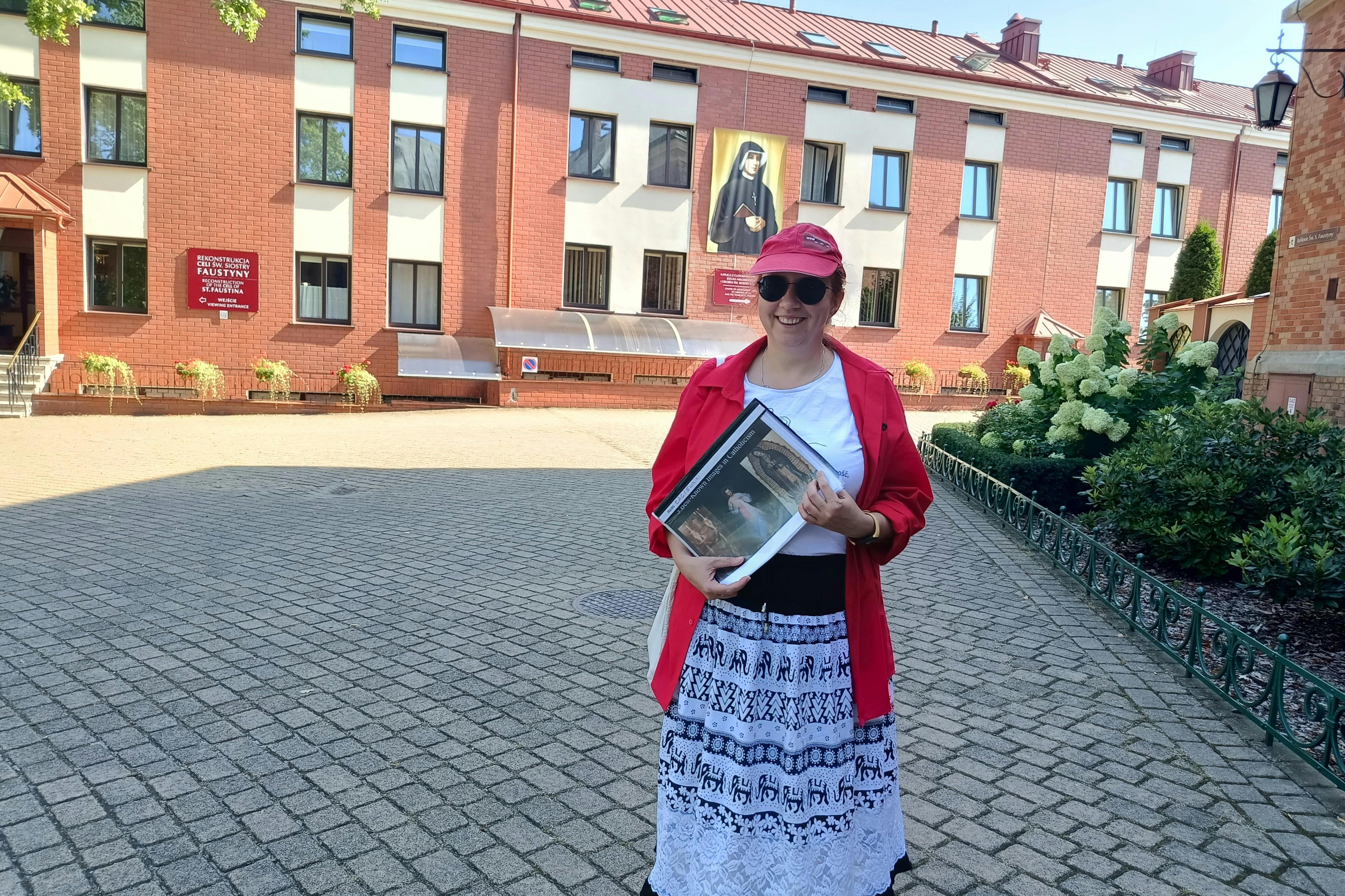 Una donna con berretto e camicia rossa tiene una cartella all'aperto, con un edificio in mattoni e un dipinto religioso sullo sfondo.