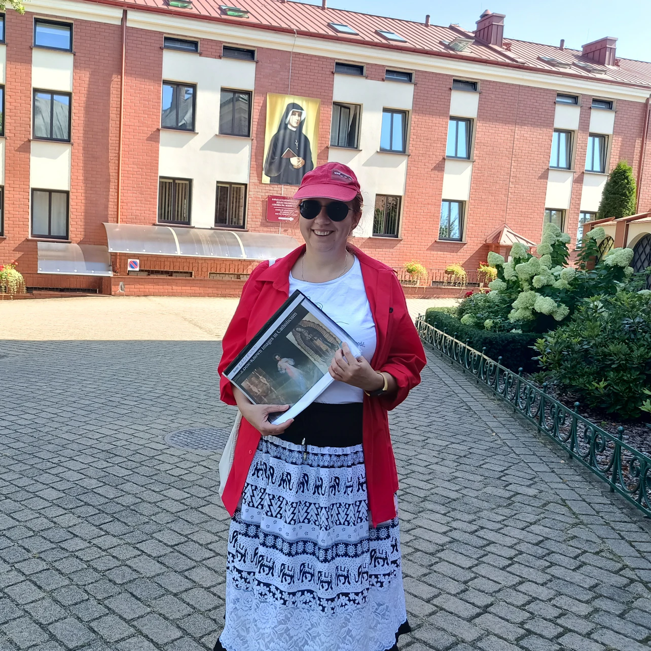 A woman in a red cap and shirt holds a folder outdoors, with a brick building and a religious painting in the background.