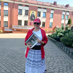 A woman in a red cap and shirt holds a folder outdoors, with a brick building and a religious painting in the background.