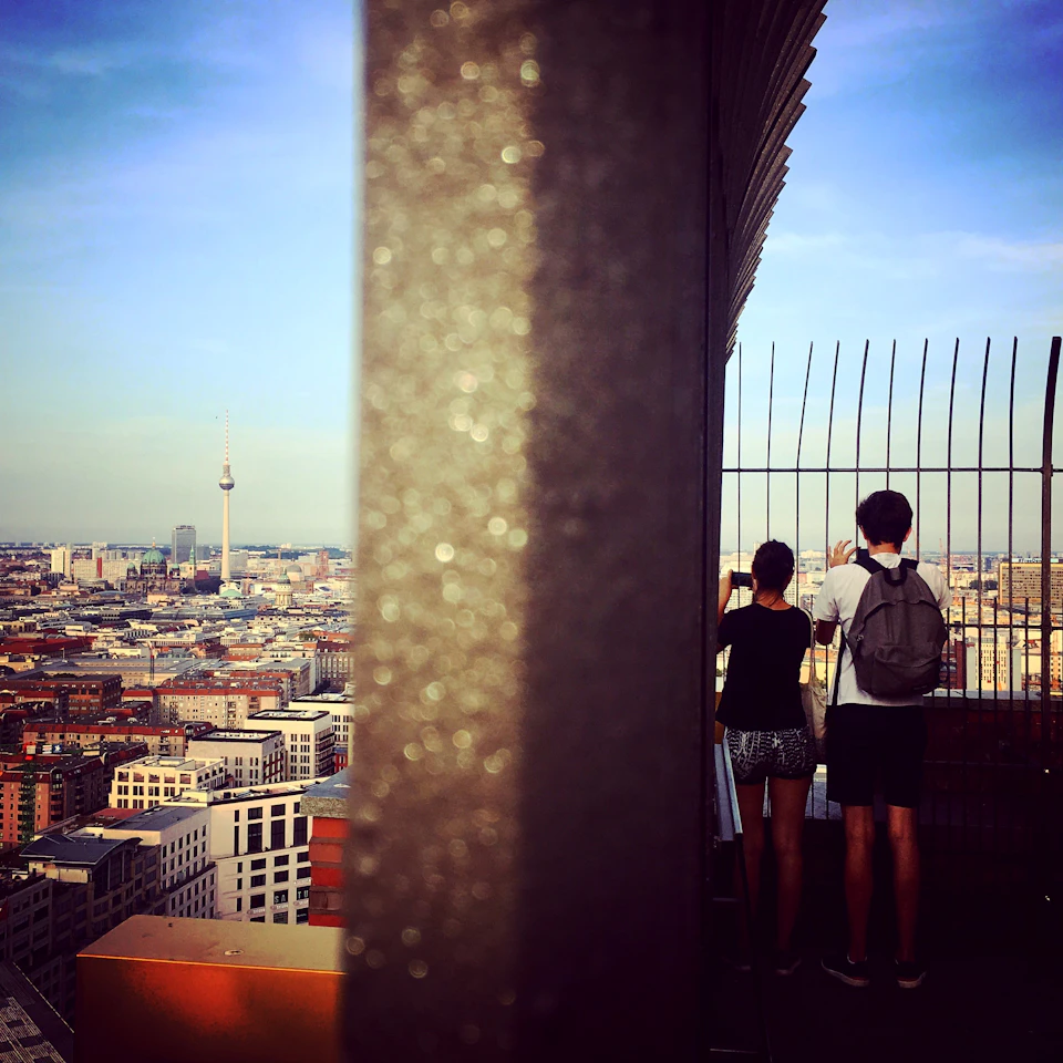 Two people stand at a railing on a rooftop observation deck, overlooking a cityscape with a tall tower in the distance.