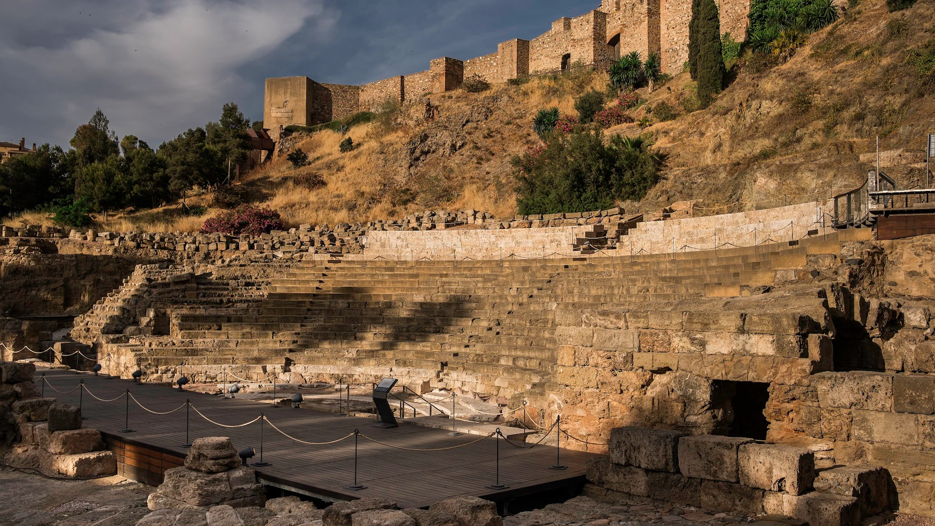 Ancient stone amphitheater with tiered seating, surrounded by ruins and vegetation under a partly cloudy sky.