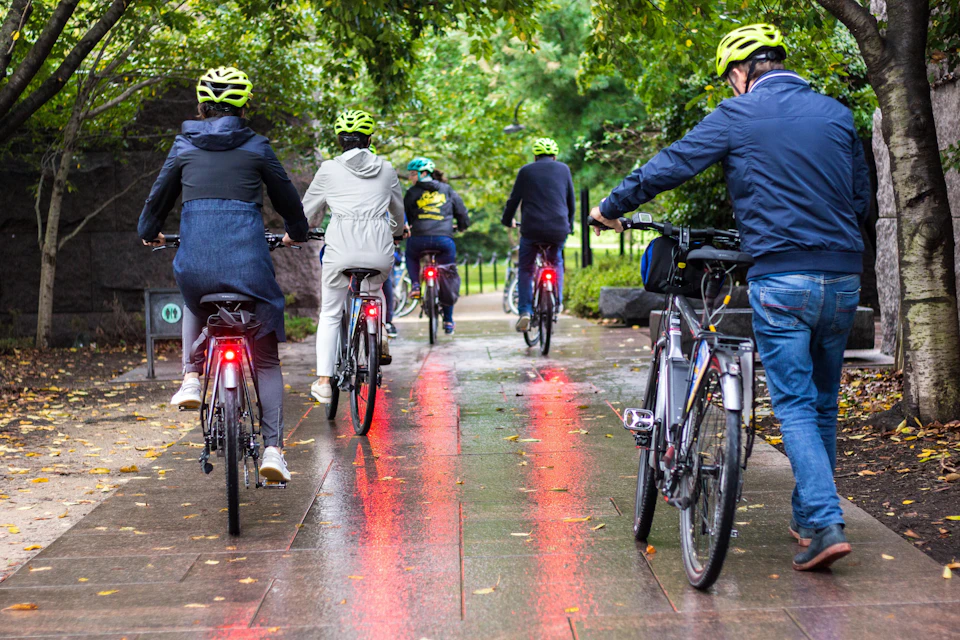 Cyclists with bright green helmets ride on a wet path through a park, trees overhead, and reflections from bike lights on the ground.