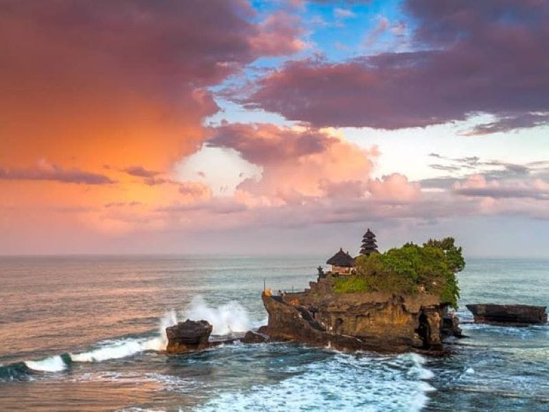 Île rocheuse avec temple entourée de vagues de l'océan, sous un ciel coloré de nuages au coucher du soleil.