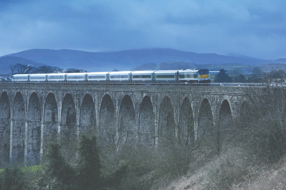 A train crossing a stone viaduct with arches on a cloudy day, with hills in the background and trees in the foreground.