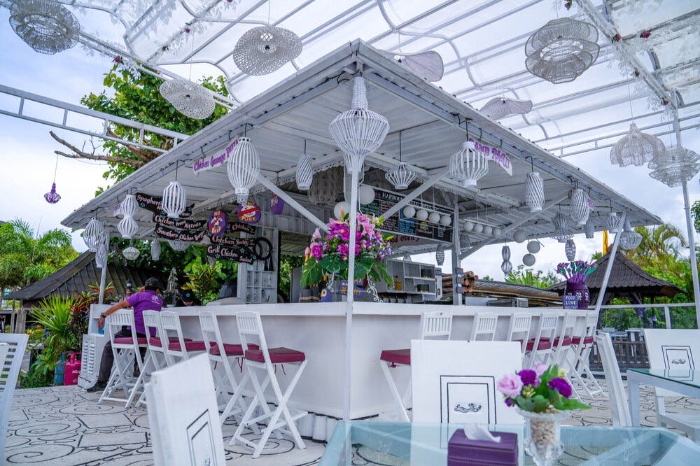 White outdoor café with a central bar, hanging lanterns, white chairs with purple cushions, and floral arrangements.