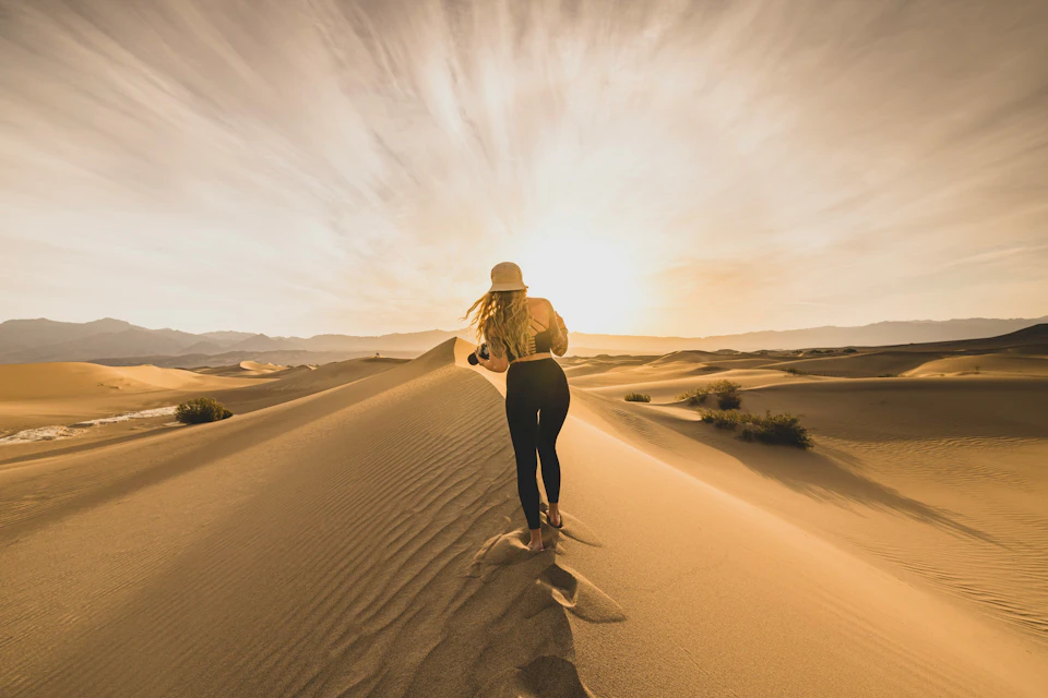 A person in a hat walking on sand dunes during sunset under a dramatic sky, with footprints trailing behind.