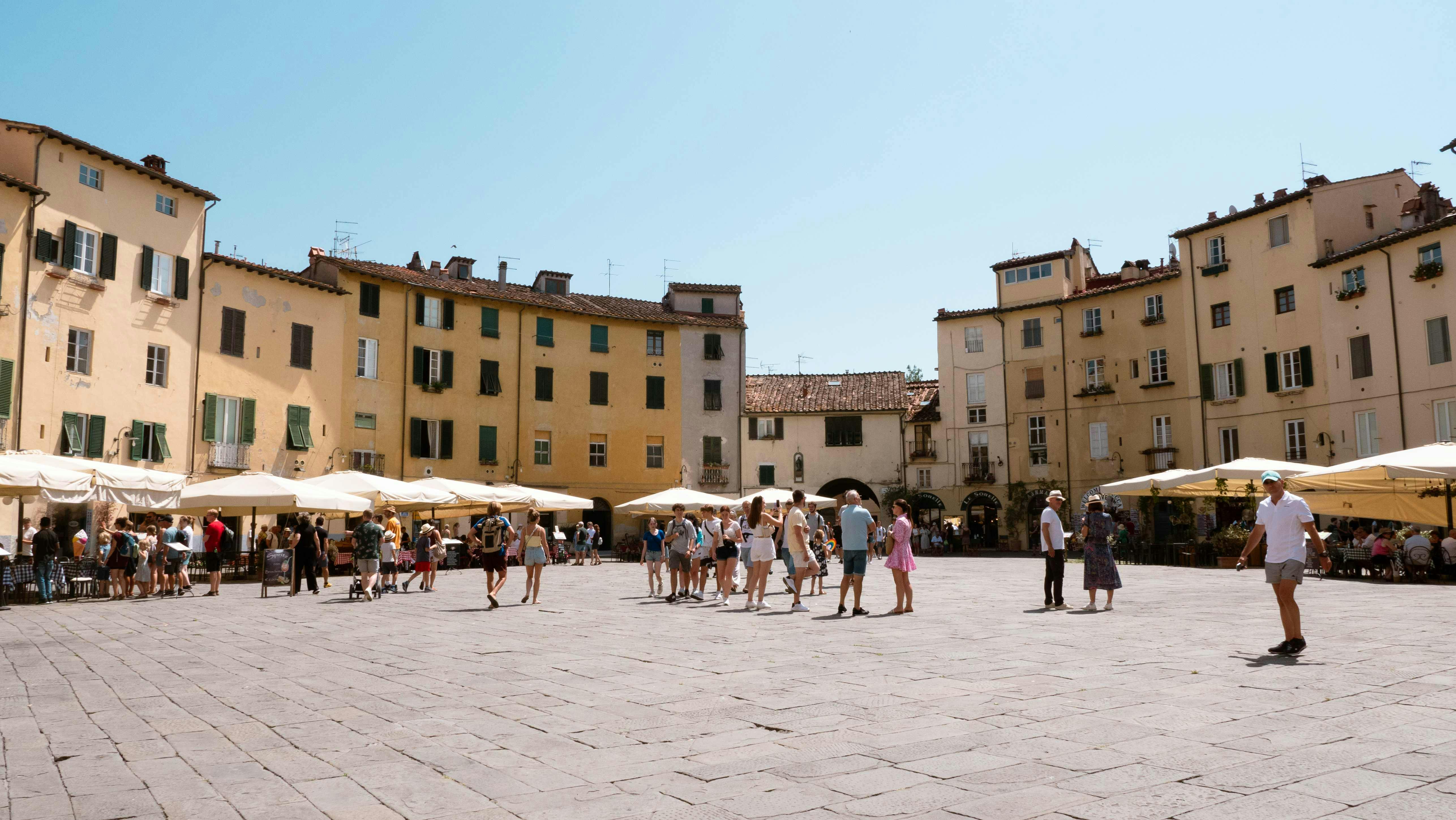 A sunlit town square with groups of people walking and relaxing near yellow and beige buildings with green shutters and outdoor umbrellas.