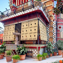 Färgglad mosaikbyggnad med utsmyckade geometriska fönster, en stor fläkt och många krukväxter framför.