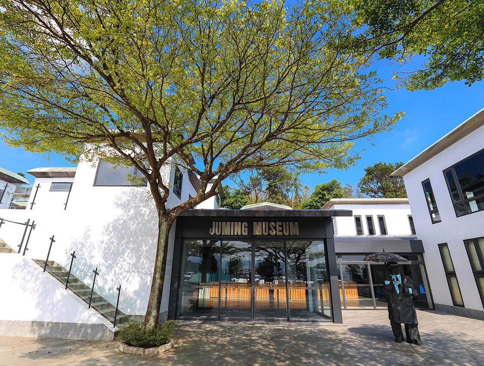 Entrance of the Juming Museum with a tree, modern buildings, and a sculpture under a clear blue sky.