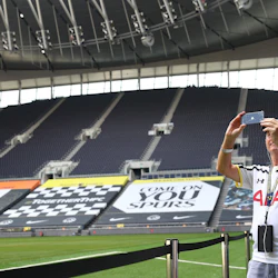 En man som bär en fotbollströja tar ett foto inuti en stor, tom stadion med "COME ON YOU SPURS" -banderoller.