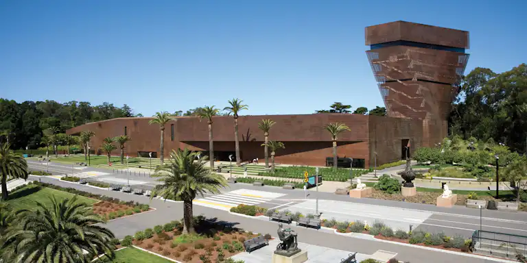 Large, modern brown building with a unique tower, surrounded by palm trees, green lawns, benches, and statues under a clear blue sky.