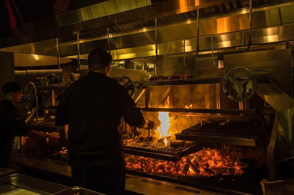 A chef grills food over an open flame in a professional kitchen with metal equipment and glowing embers.