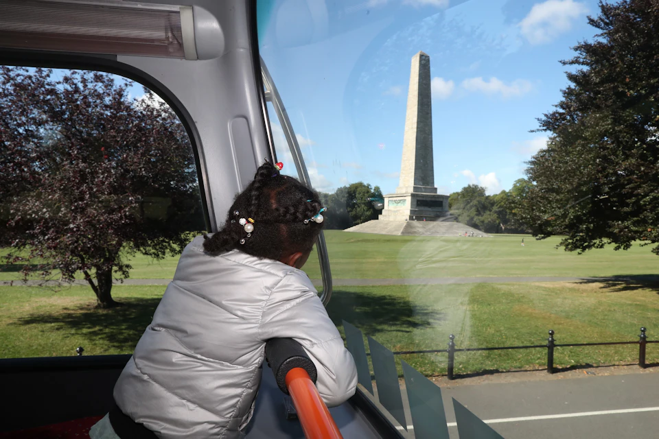 Child in a silver jacket looks out of a bus window at a tall stone obelisk monument in a park under a blue sky.
