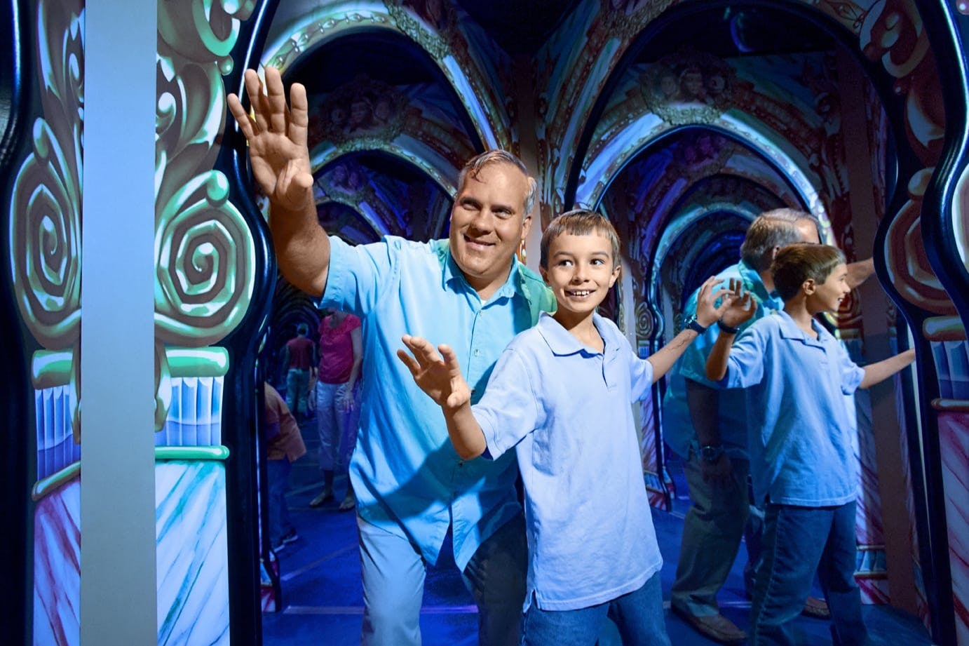 Un homme et un garçon explorent un labyrinthe coloré recouvert de miroirs, souriants, tendant la main pour toucher les surfaces réfléchissantes.