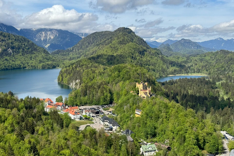 A scenic view of a castle on a forested hill, a small village, lakes, and mountains in the background under a partly cloudy sky.