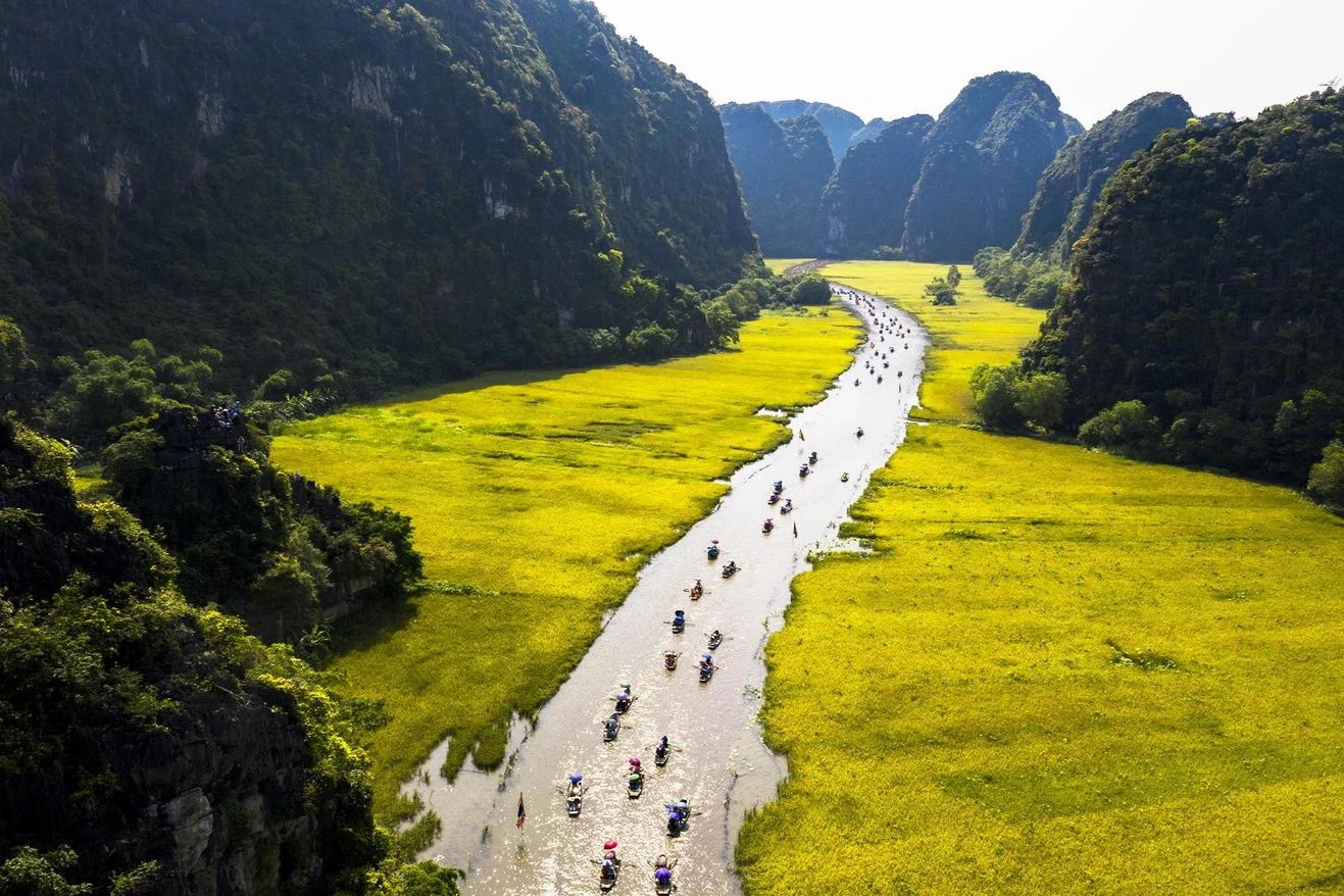 A river winding through vibrant green fields and steep hills, with numerous small boats carrying people down the water.