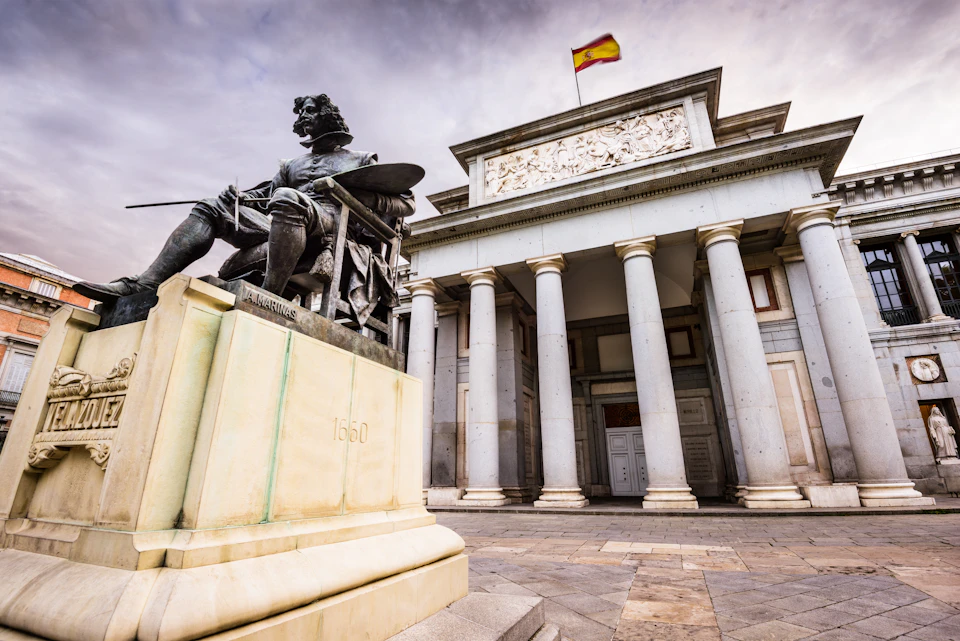 Statue of a seated figure in front of a grand building with columns and a Spanish flag on the roof. Overcast sky.