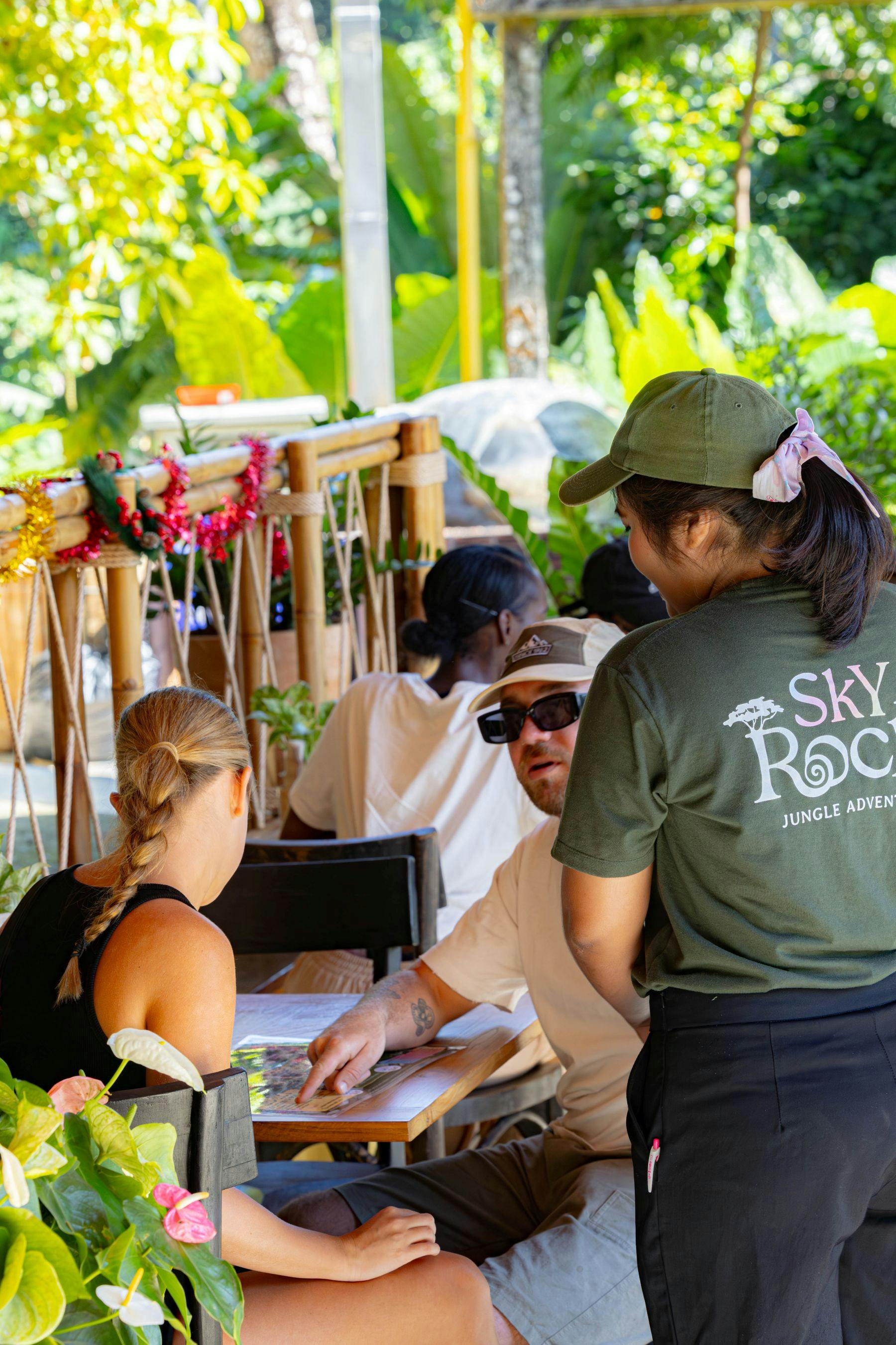 A group of people interacting at an outdoor table surrounded by plants and bamboo decor. One person points at a map.