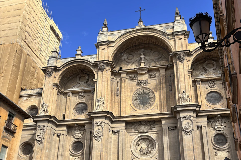 Ornate façade of a historic building with arches, detailed carvings, round windows, and a cross on top under a clear blue sky.