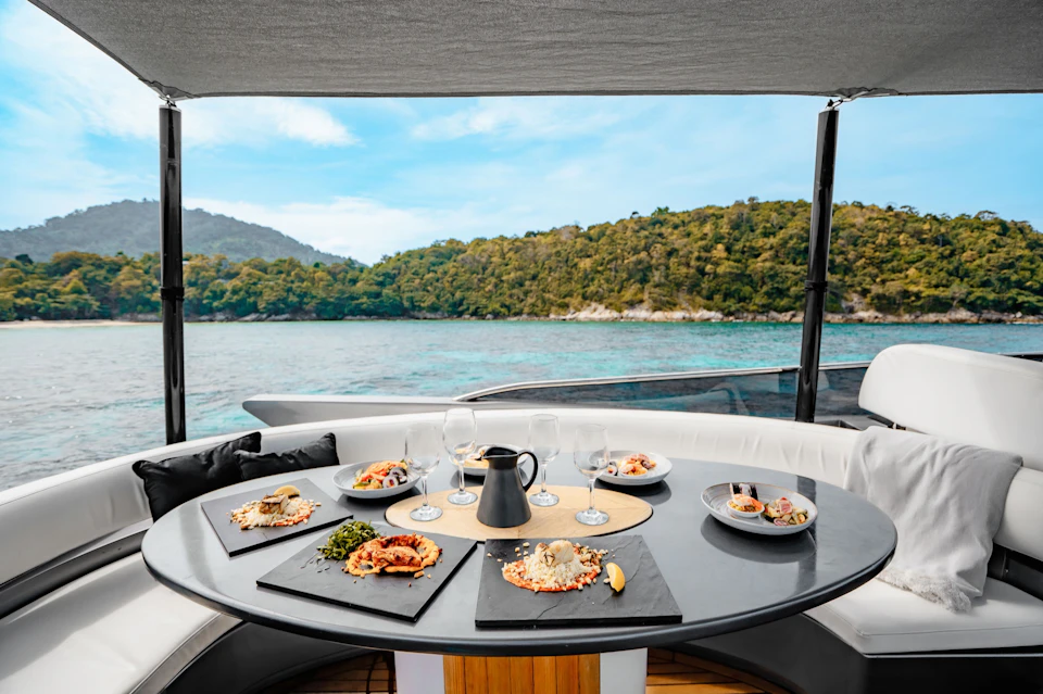 A table on a boat set with dishes of food, wine glasses, and a pitcher, with a view of the ocean and a forested hillside.