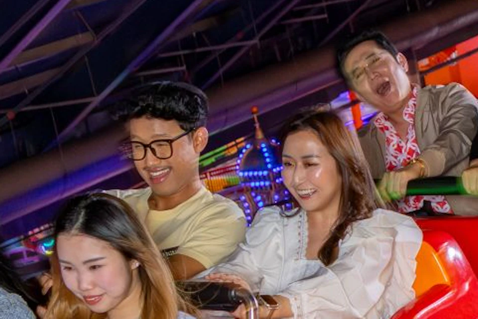 A group of people riding a brightly colored roller coaster indoors with visible structural beams and lighting.