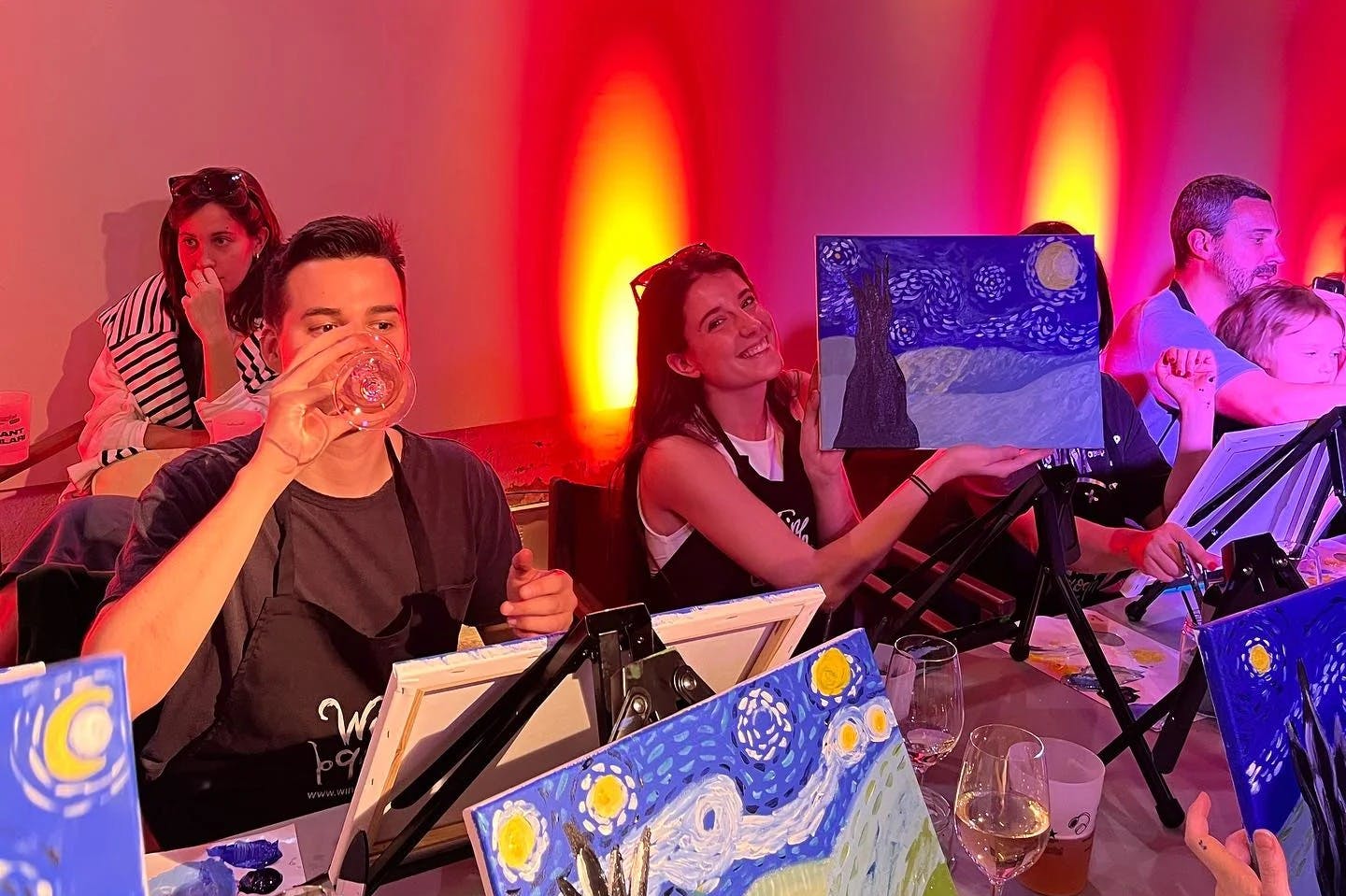 People painting Van Gogh's Starry Night replicas at a table. One person sips a drink, while another smiles and shows their painting.
