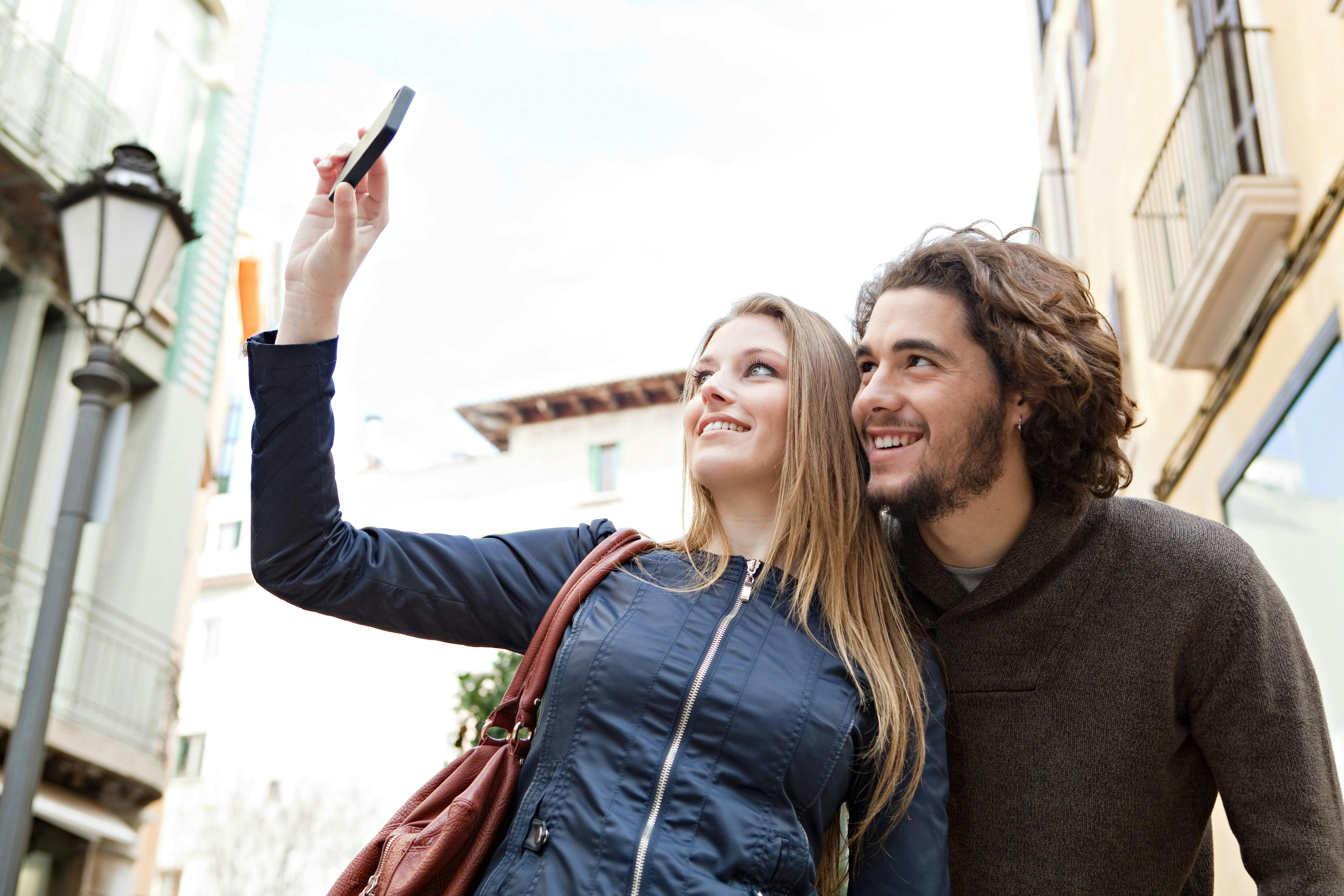 Two people smiling and taking a selfie outdoors near buildings with balconies.