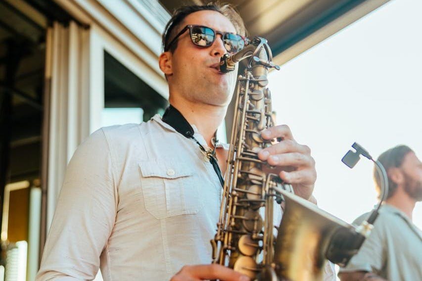 Man som spelar saxofon, bär solglasögon och en lätt skjorta. En annan person i bakgrunden under en takkonstruktion.