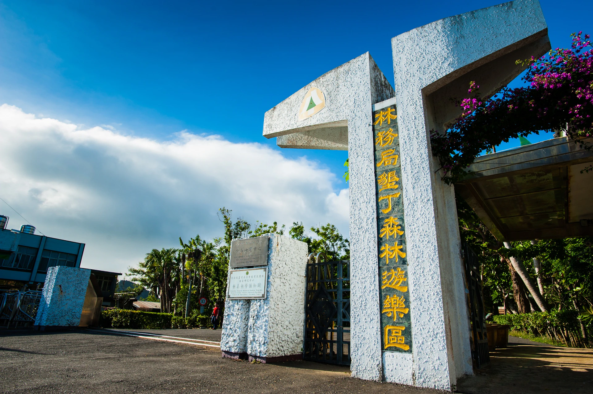 Entrance gate to a park with yellow text on a white structure, surrounded by greenery under a clear blue sky.