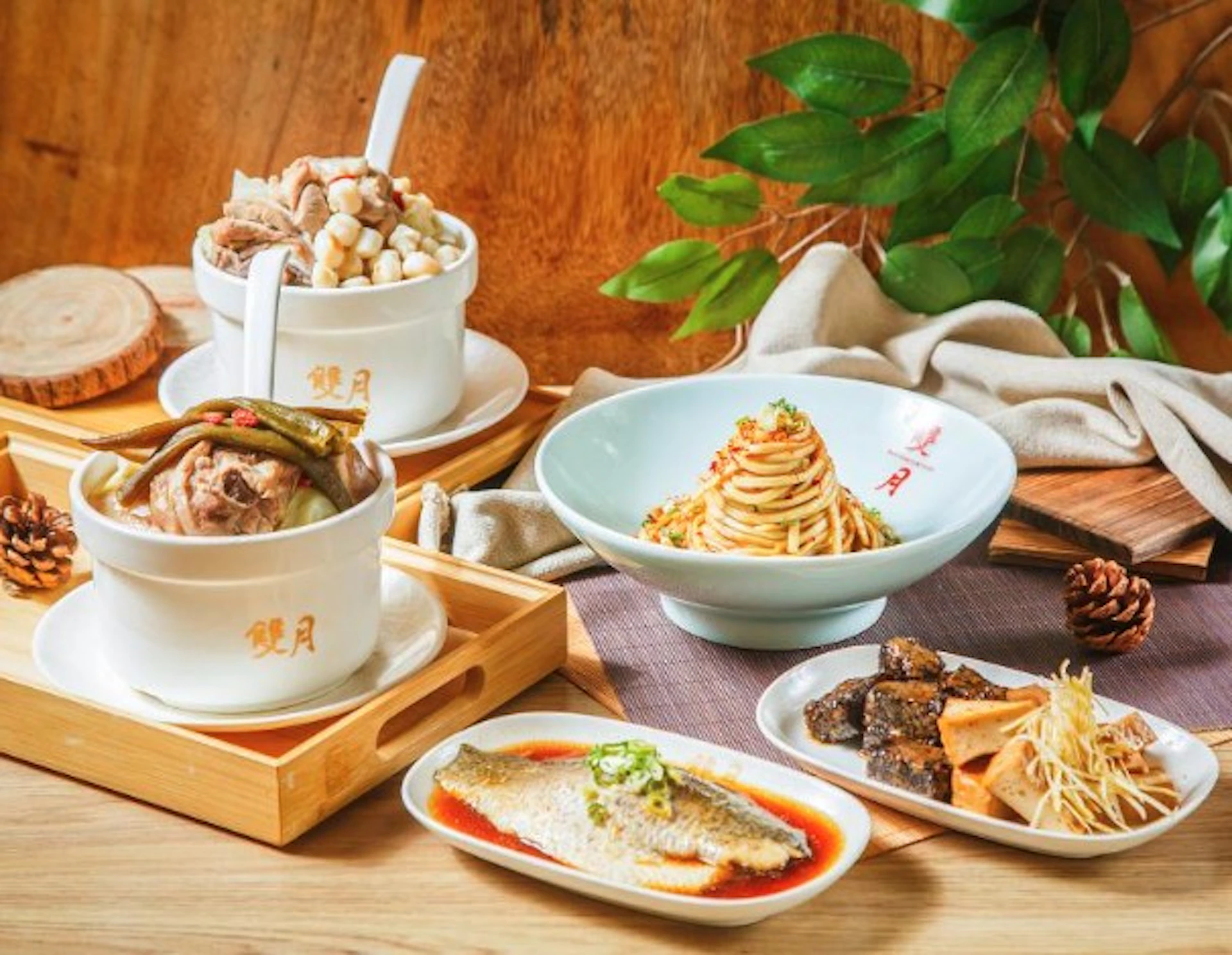 A variety of Chinese dishes arranged on a table, including soup, noodles, fish, and side items, with green leaves in the background.