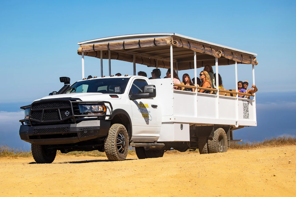 Open-air tour truck with a canopy, carrying seated passengers on a sunny day in a sandy, outdoor area.