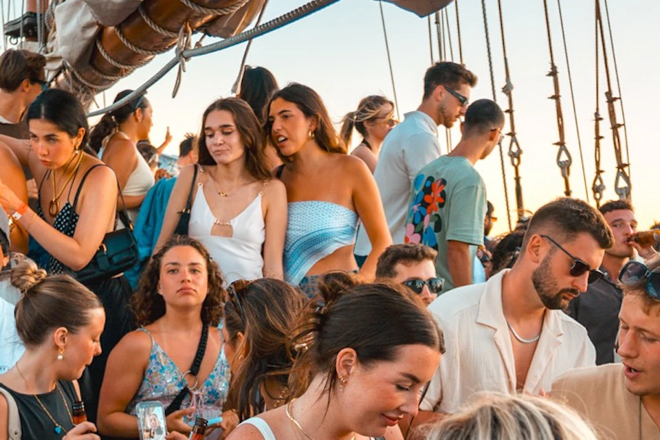 A group of people enjoying a party on a boat during sunset, with a DJ performing in the foreground and sails in the background.