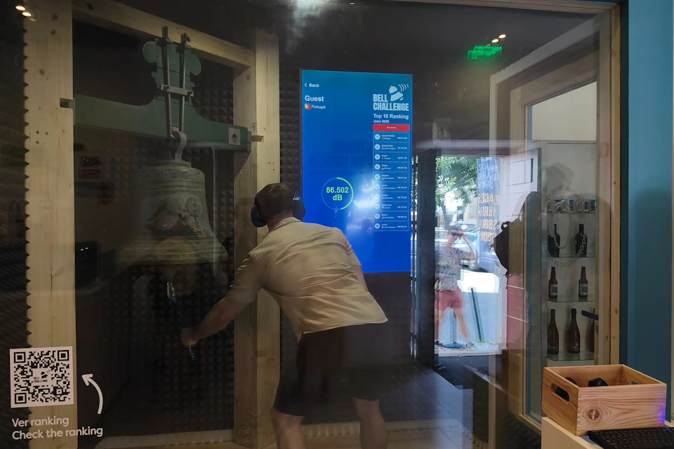 A person rings a large bell in a soundproofed room with a blue leaderboard display showing a sound level score.