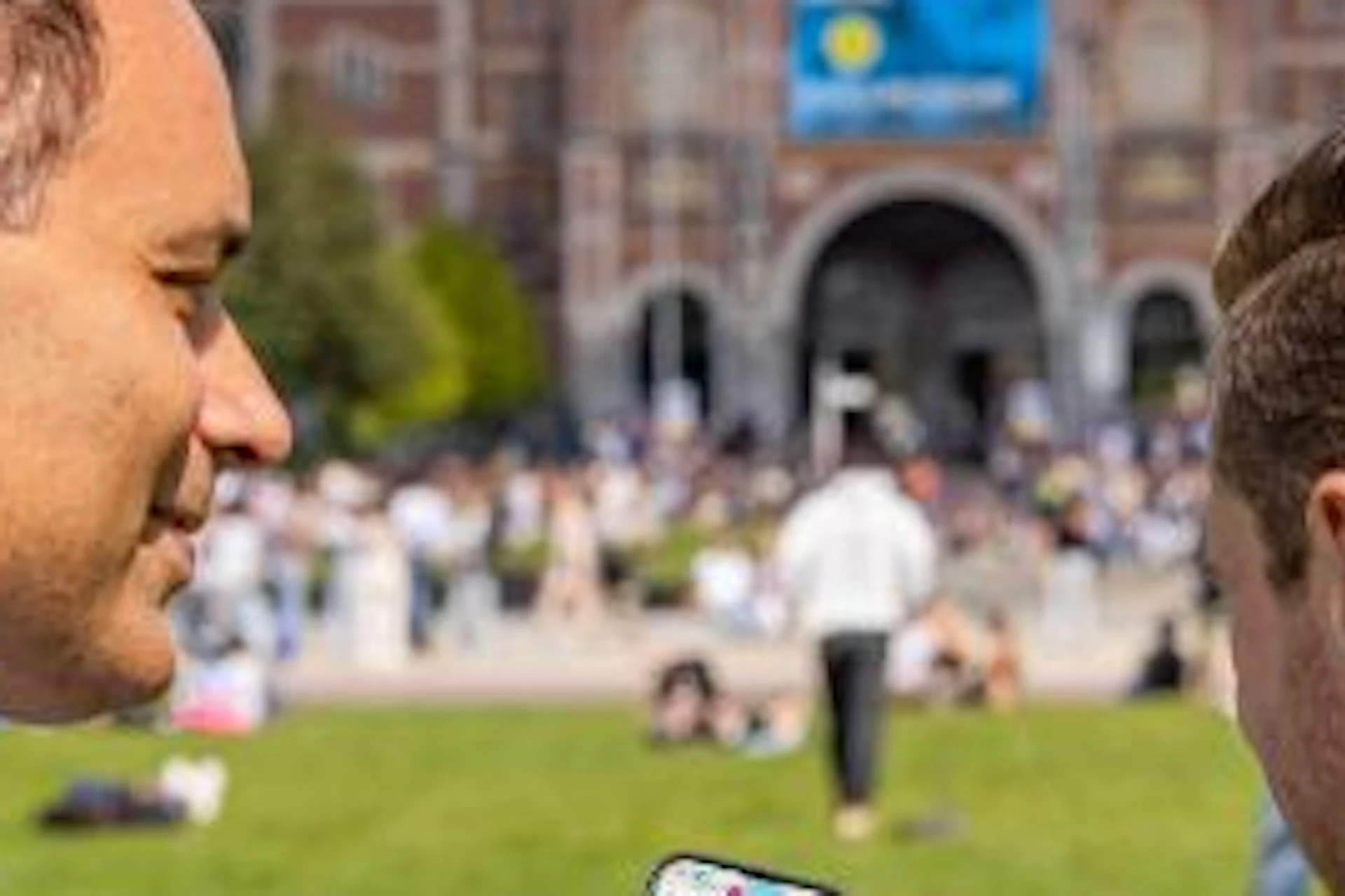 Two men stand in a park, one holding a smartphone displaying a map. A large building with an archway is in the background.