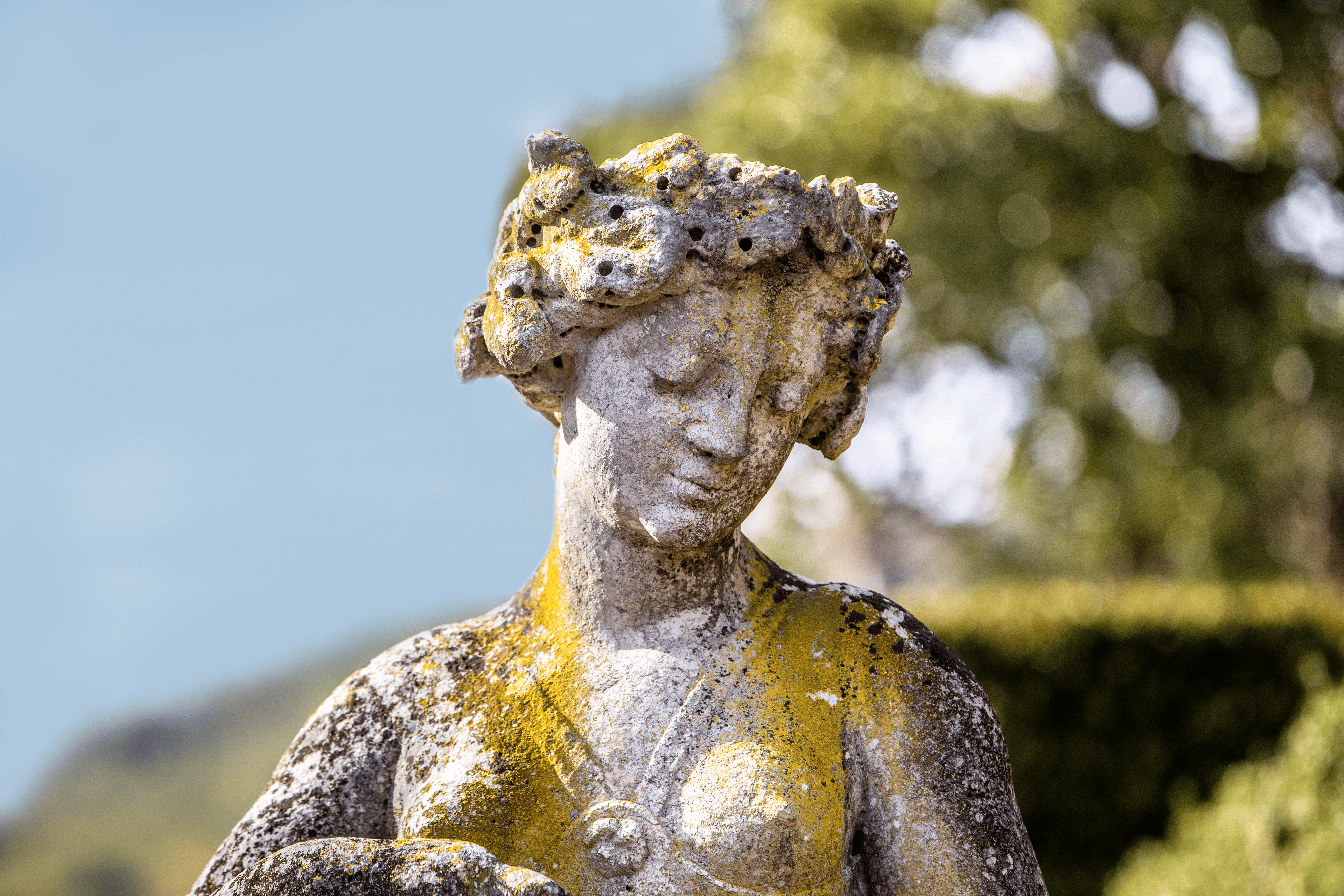 Statua in pietra danneggiata dalle intemperie di una donna dall'espressione serena, ricoperta da macchie di muschio. Alberi e cielo sullo sfondo sfocato.