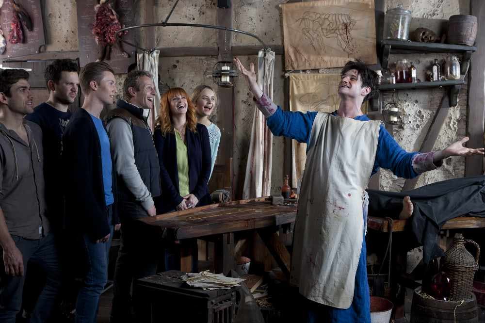 A group of five people laughing and watching a man in an apron performing in a rustic, art-studio-like setting.