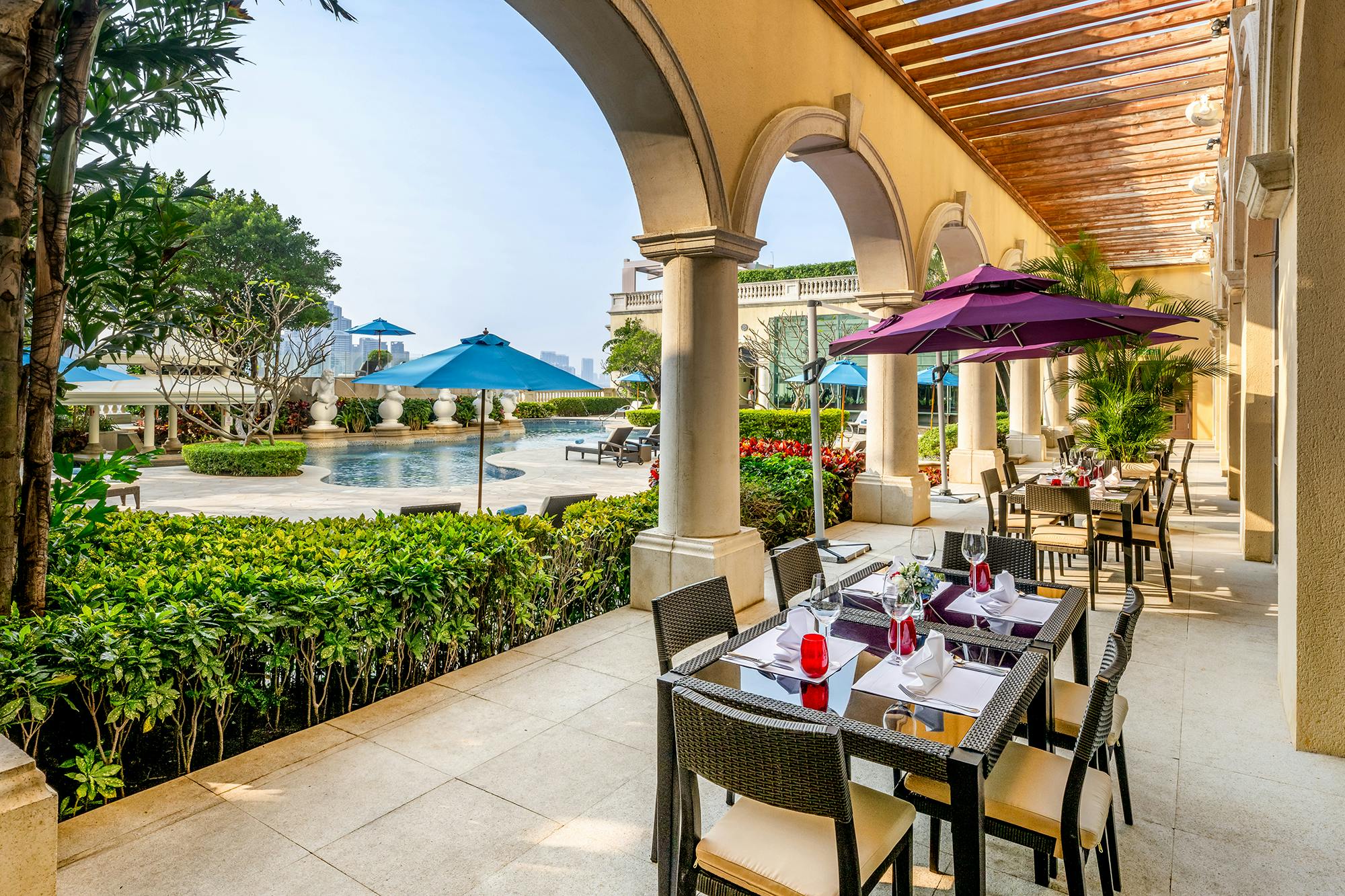 Outdoor dining area with tables and chairs under colorful umbrellas, near a pool surrounded by greenery and arches.