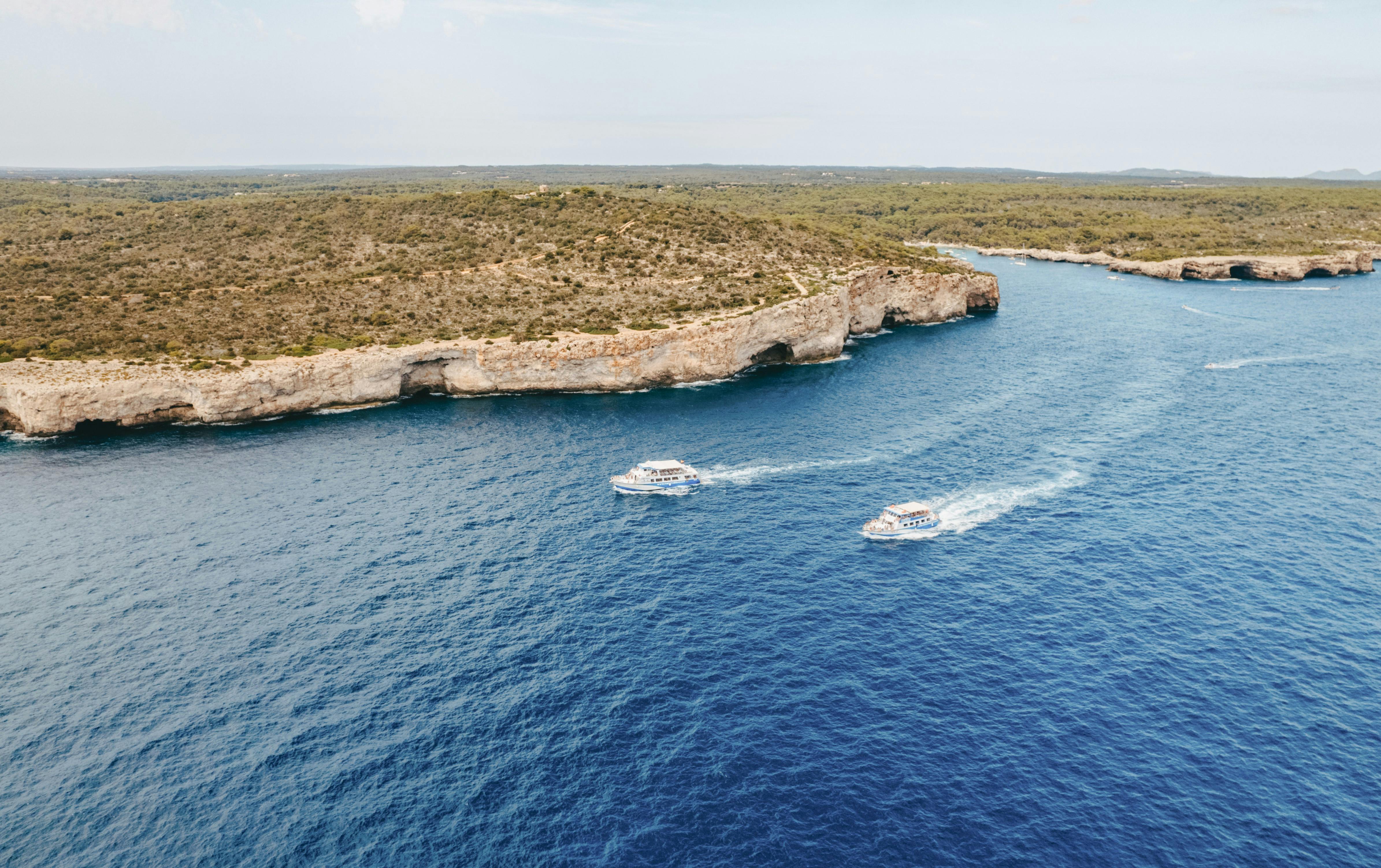 Interior view of a ferry with beige seats and tables. Large windows show a calm sea and sailboats outside.