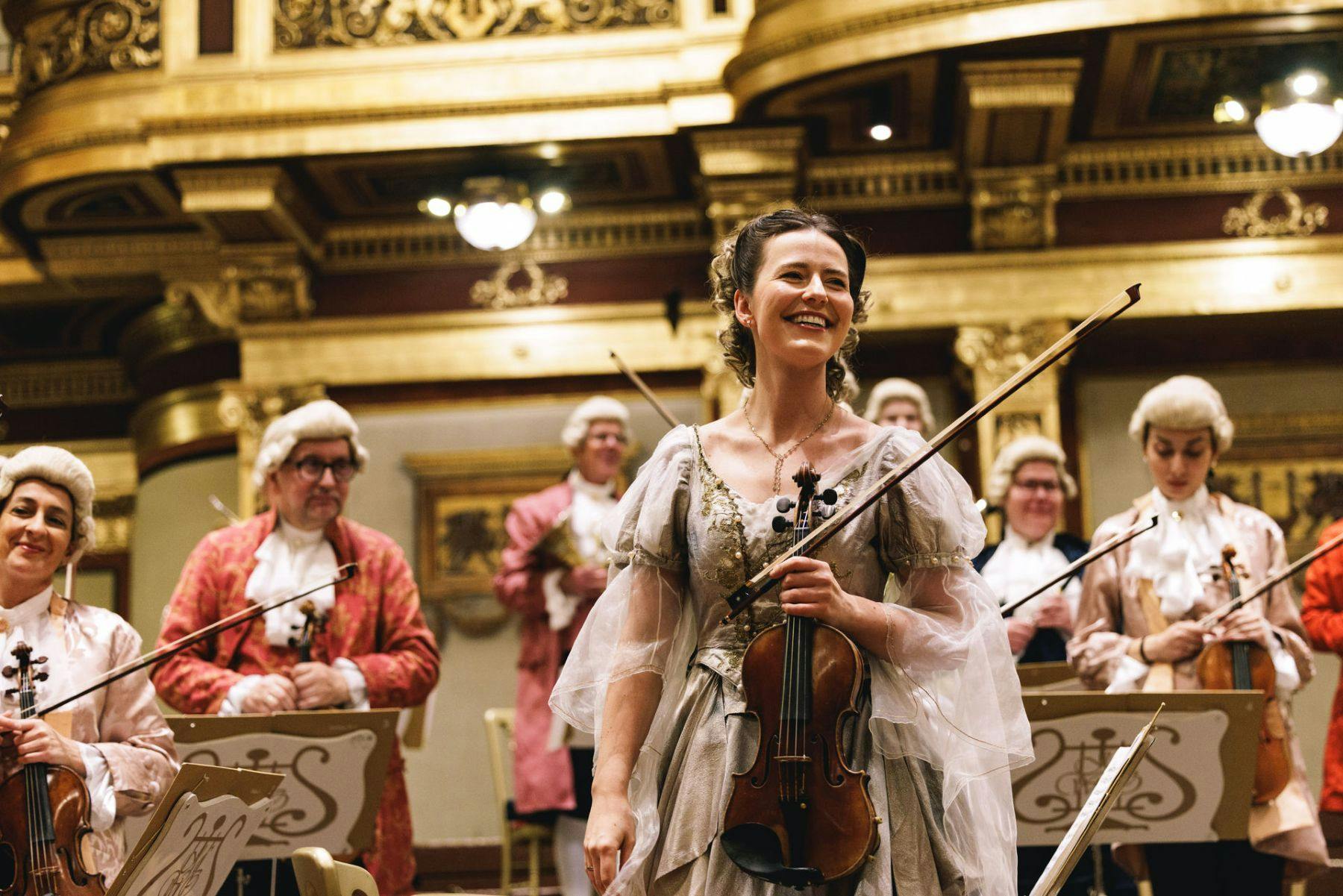 A smiling woman in period costume holds a violin on stage, surrounded by other musicians in a lavishly decorated hall.