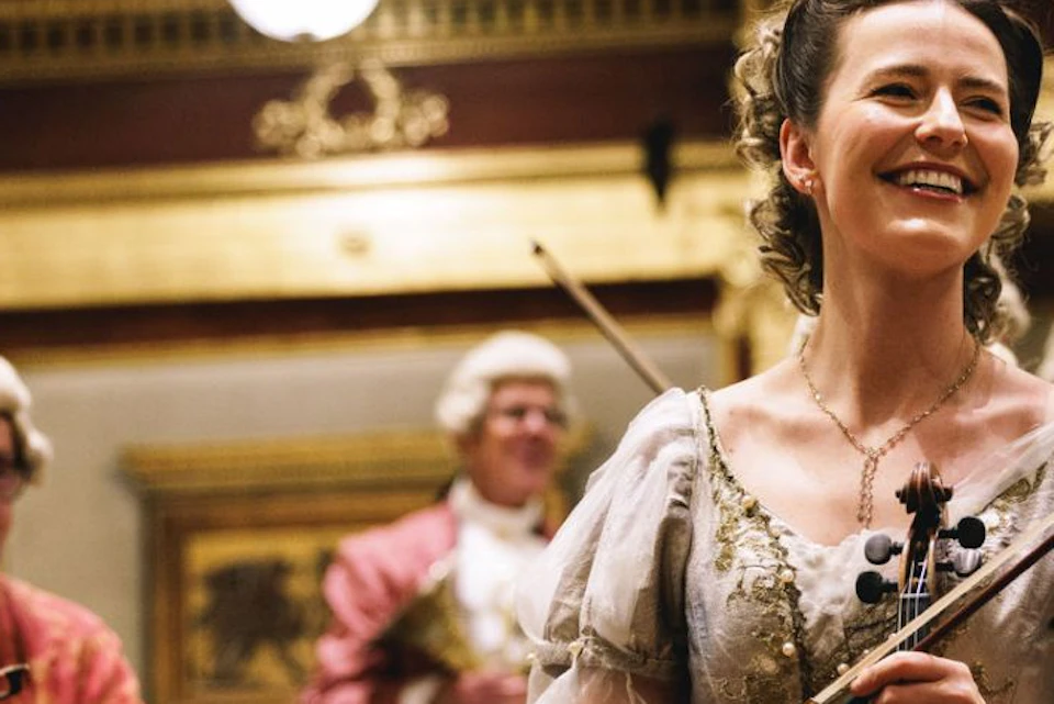 A smiling woman in period costume holds a violin on stage, surrounded by other musicians in a lavishly decorated hall.