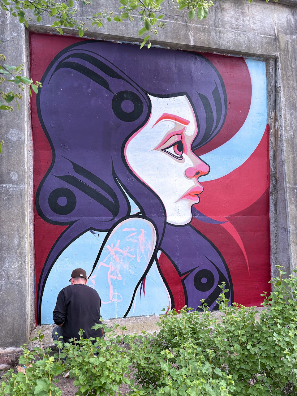 A person is painting a large mural of a stylized female profile with purple hair on a concrete wall, surrounded by greenery.