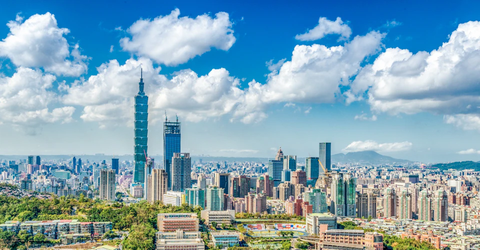 A city skyline with the Taipei 101 skyscraper, surrounded by modern buildings under a blue sky with scattered clouds.