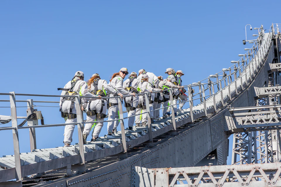 A group of people in climbing gear and helmets walks up an inclined metal structure on a clear day.