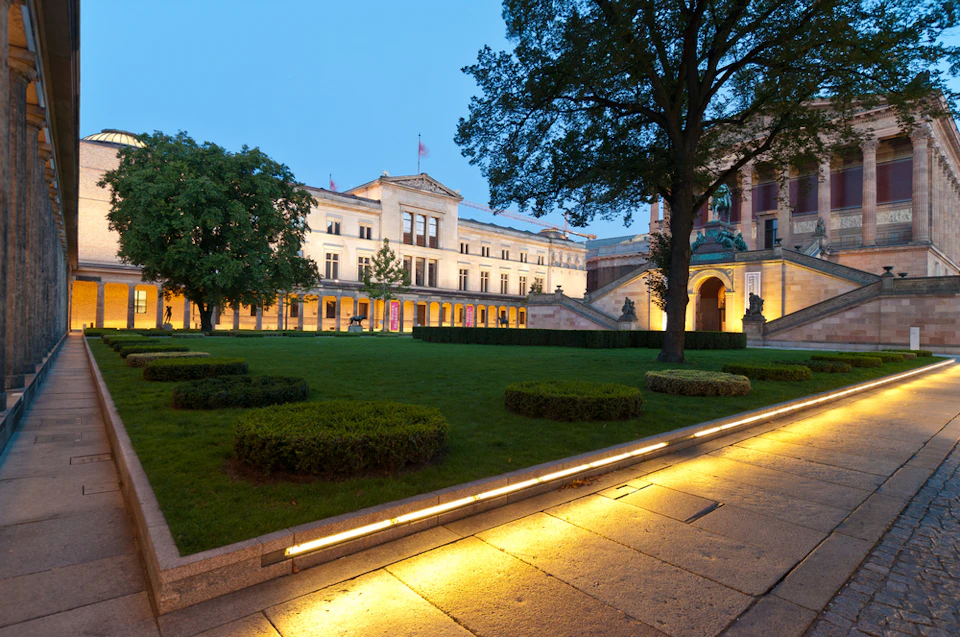 Illuminated courtyard with a trimmed lawn, bushes, trees, and a grand building in the background at dusk.