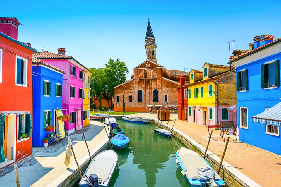 A canal lined with colorful houses, small boats on the water, and a church with a tall steeple in the background.