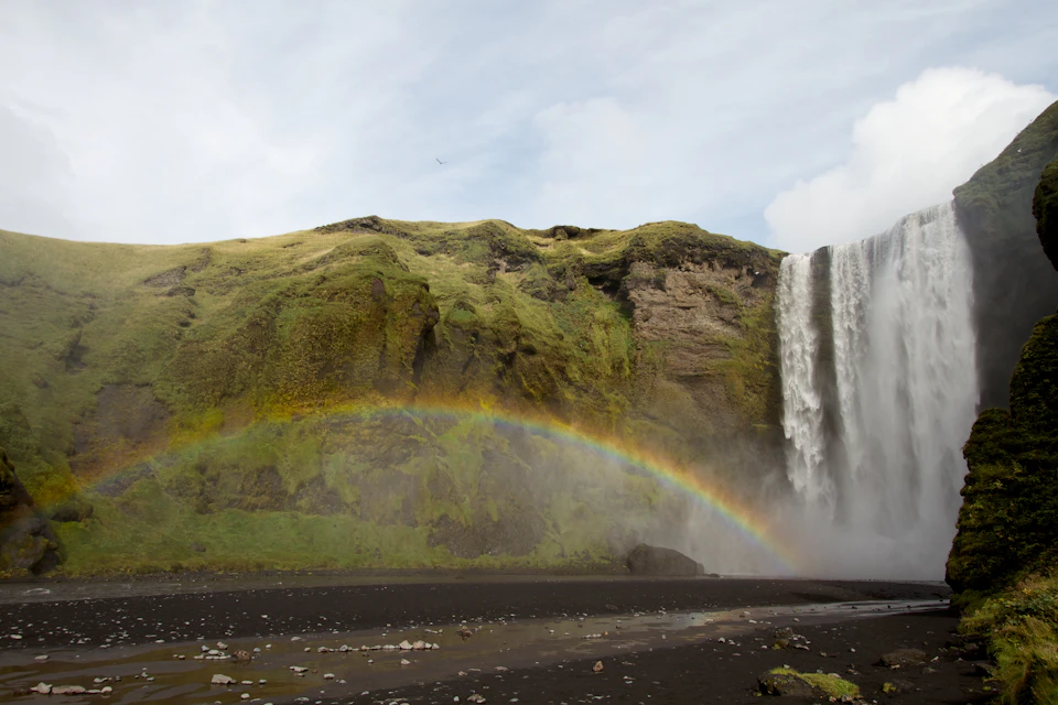 Tall waterfall cascading down a green cliff with a clear, arched rainbow forming at its base.