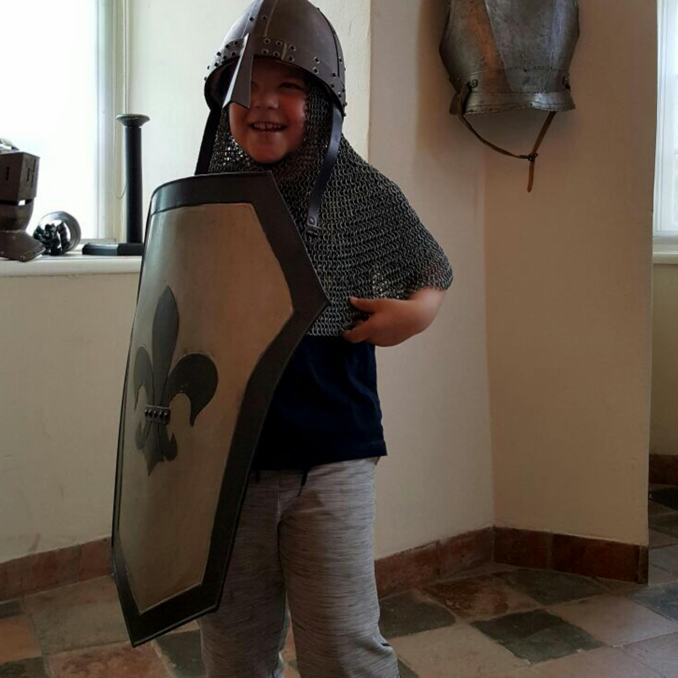 Ένα παιδί που φοράει αλυσιδωτή αλυσίδα και κράνος κρατά μια ασπίδα με fleur-de-lis, που στέκεται σε ένα δωμάτιο με μεσαιωνικές οθόνες πανοπλίας.